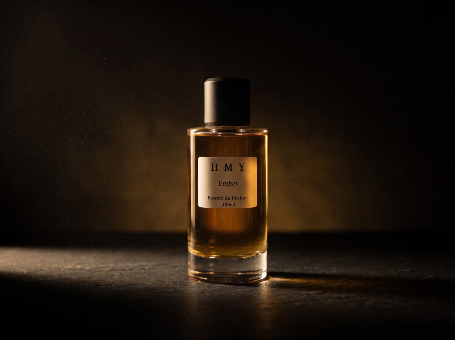 The HMY Ember bottle in warm low light — amber extrait behind clear glass, matte black cap, the H M Y label