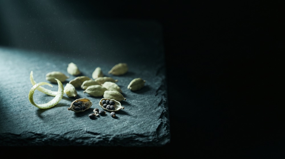 Green cardamom pods and a curl of citrus peel on dark slate in cool light