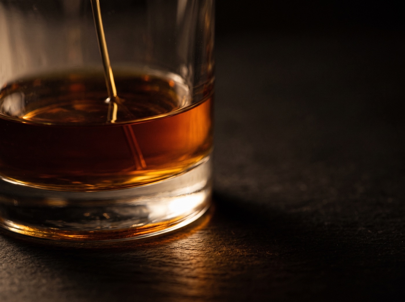 Deep amber perfume liquid inside clear glass, catching warm light against dark slate